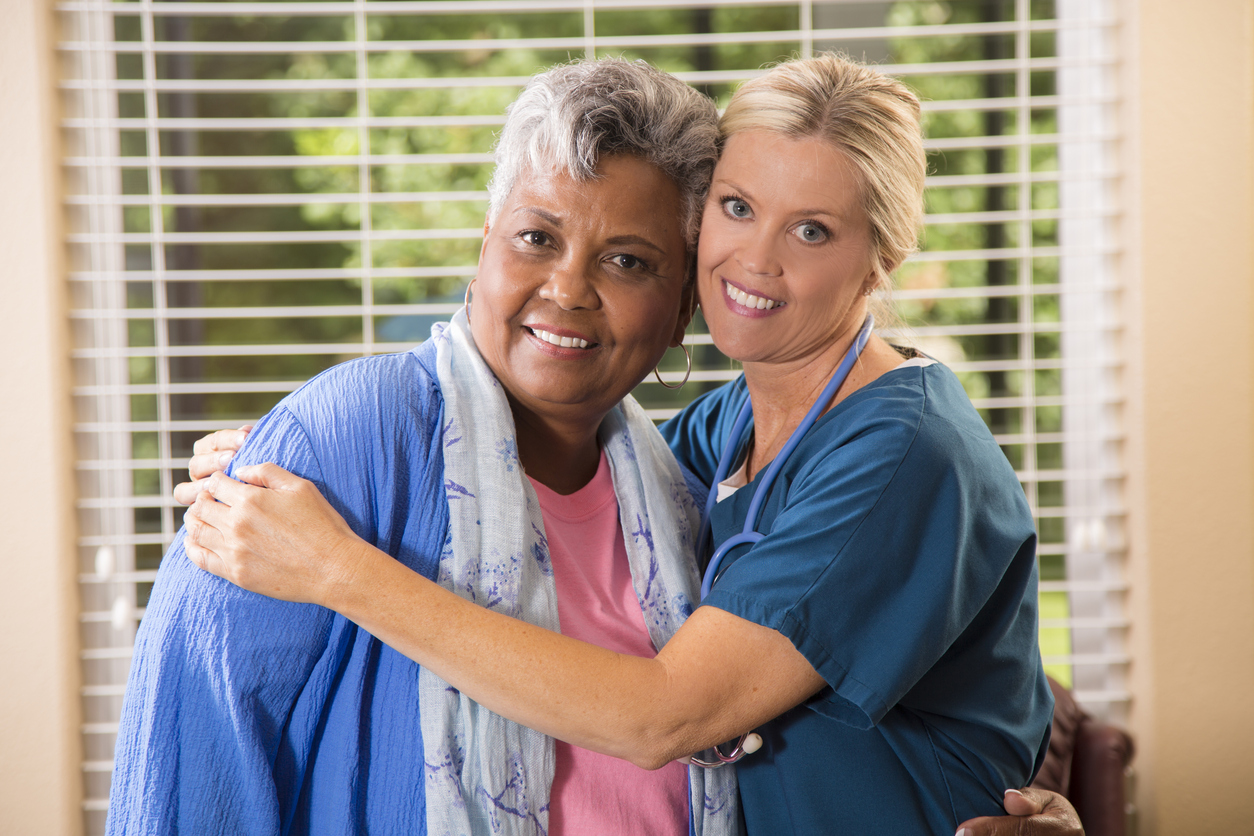 Close-up view of a caring home healthcare nurse with her senior adult female patient at home or an assisted living facility.  The female patient gets an affectionate hug from her nurse.  Backyard can be seen through the windows.